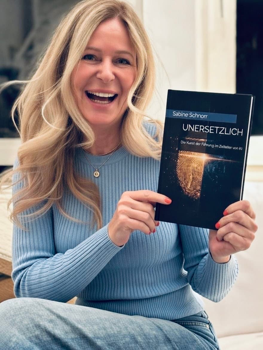Lächelnde blonde Frau im blauen Pullover präsentiert stolz ihr Buch „UNERSETZLICH“ von Sabine Schnorr.