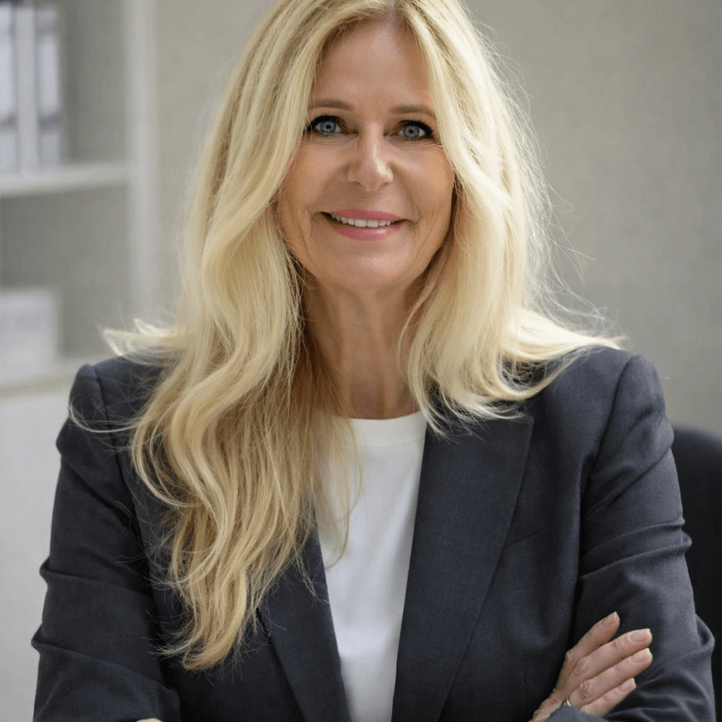 Lächelnde blonde Frau im dunklen Blazer mit verschränkten Armen blickt direkt in die Kamera.