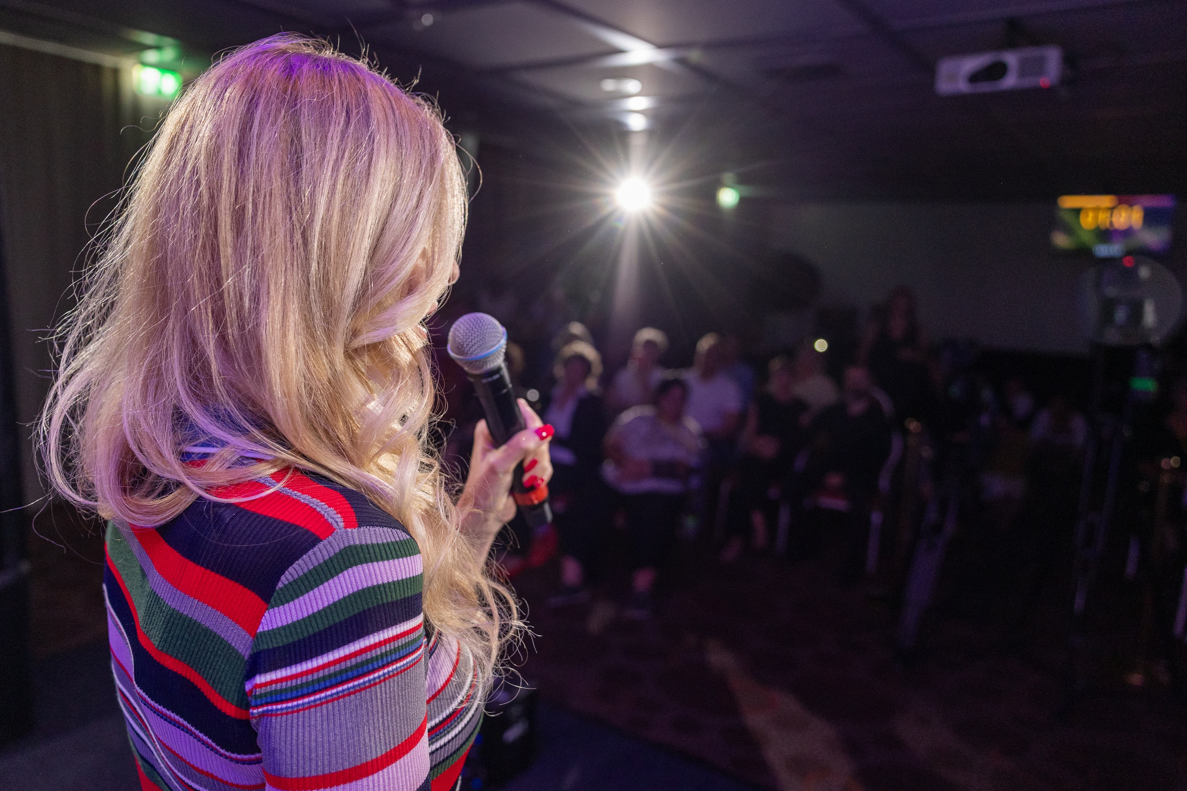 Rückansicht einer blonden Frau im bunt gestreiften Pullover, die vor Publikum in ein Mikrofon spricht.