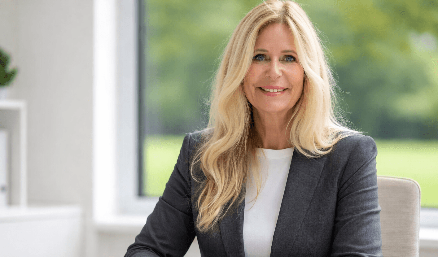 Eine lächelnde blonde Frau in einem grauen Blazer und weißem Oberteil sitzt in einem Büro.